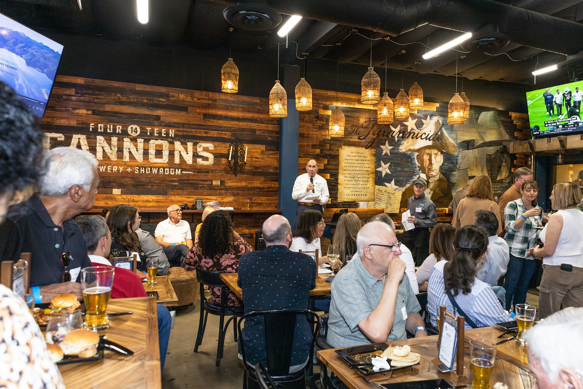 A lively gathering at a brewery, featuring a speaker, food, drinks, and an audience engaged in conversation.