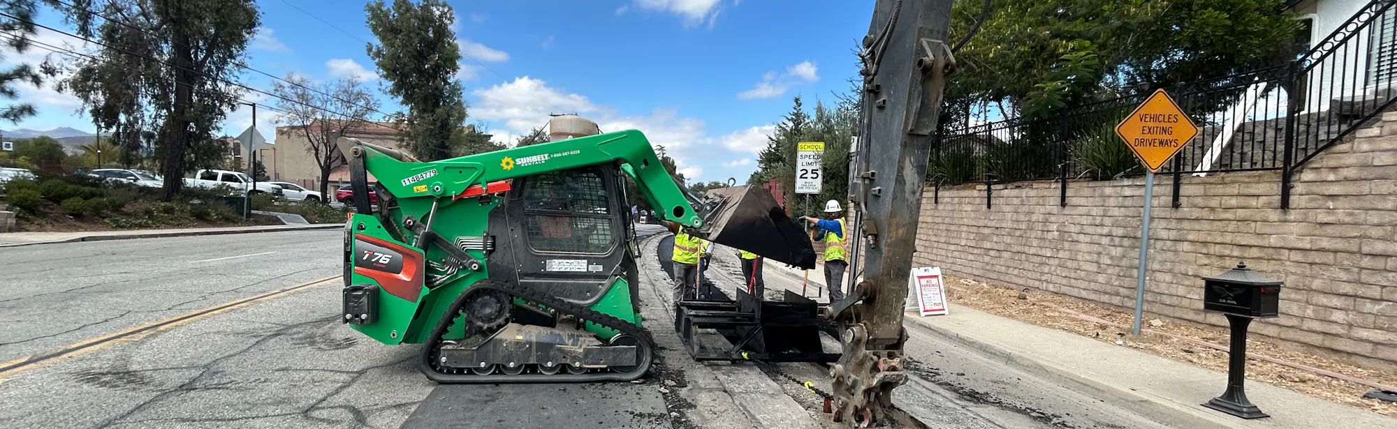 A green skid steer and an excavator are working on road maintenance, with construction signs and workers visible in the background.