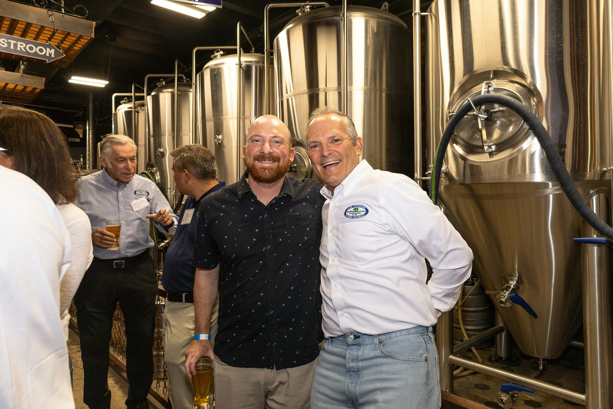 The image shows a group of people enjoying a gathering in a brewery, with stainless steel tanks in the background.