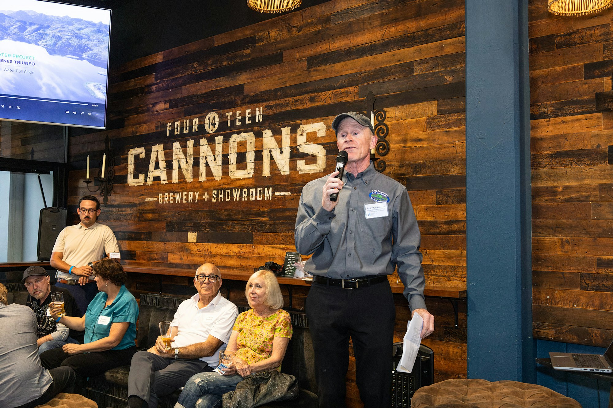 A man speaks at an event in a brewery, while attendees sit and listen. A screen displays information about a project.