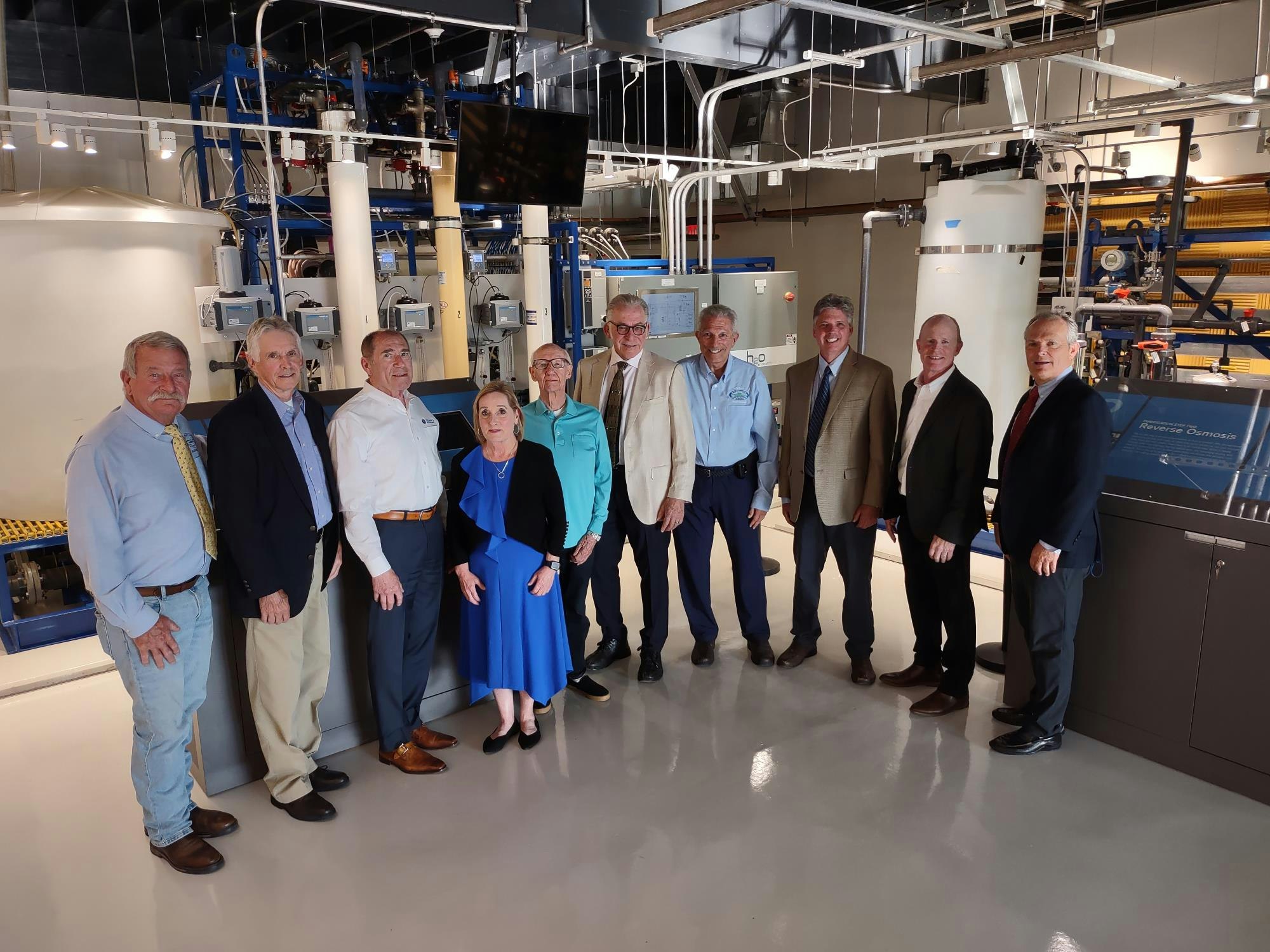A group of people are posing inside an industrial facility, with pipes and machinery in the background.