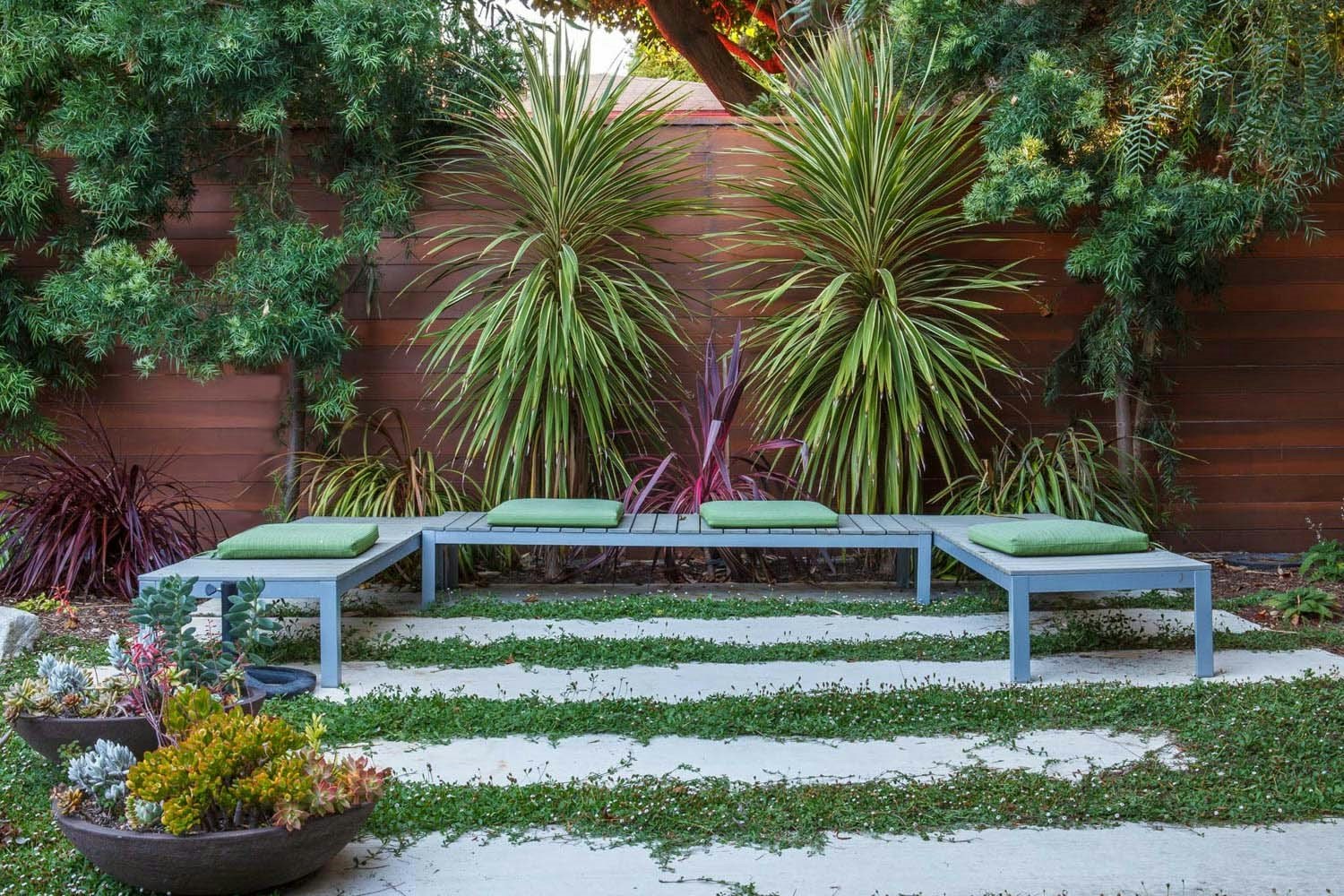 Modern garden with a wooden fence, tropical plants, a gray L-shaped bench, green cushions, and potted succulents.
