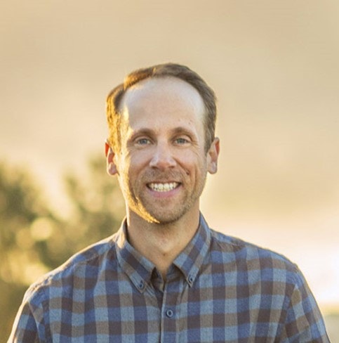 A smiling man in a plaid shirt with a natural background during golden hour.