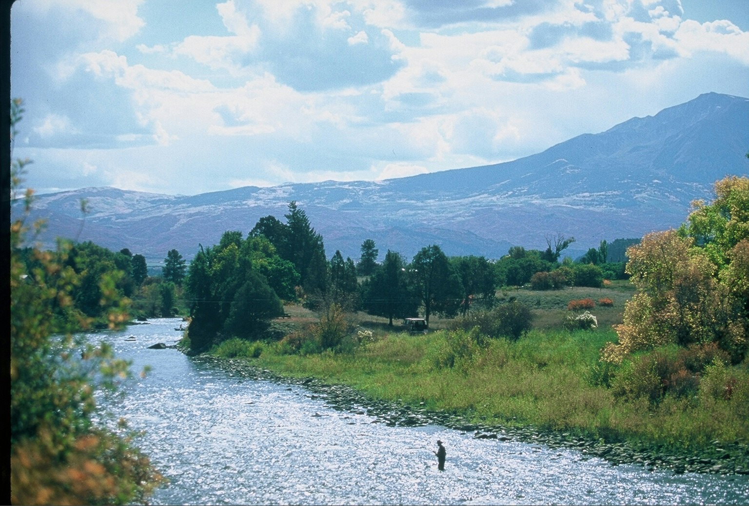 A person is fishing in a sparkling river with mountains in the background and lush greenery around.