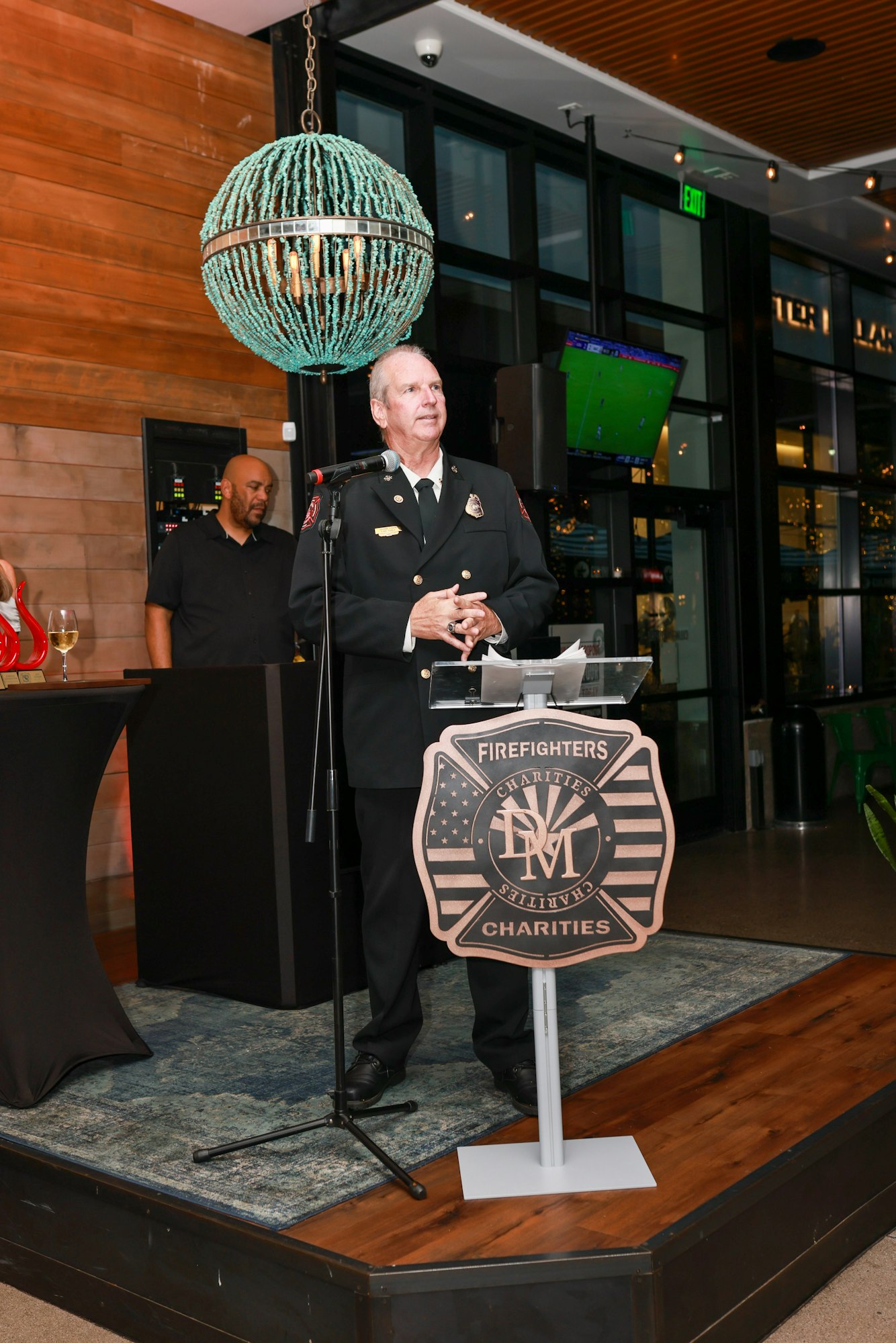 A person in a firefighter uniform speaks at a podium with "Firefighters Charities" on it, inside a venue with a chandelier and TV.