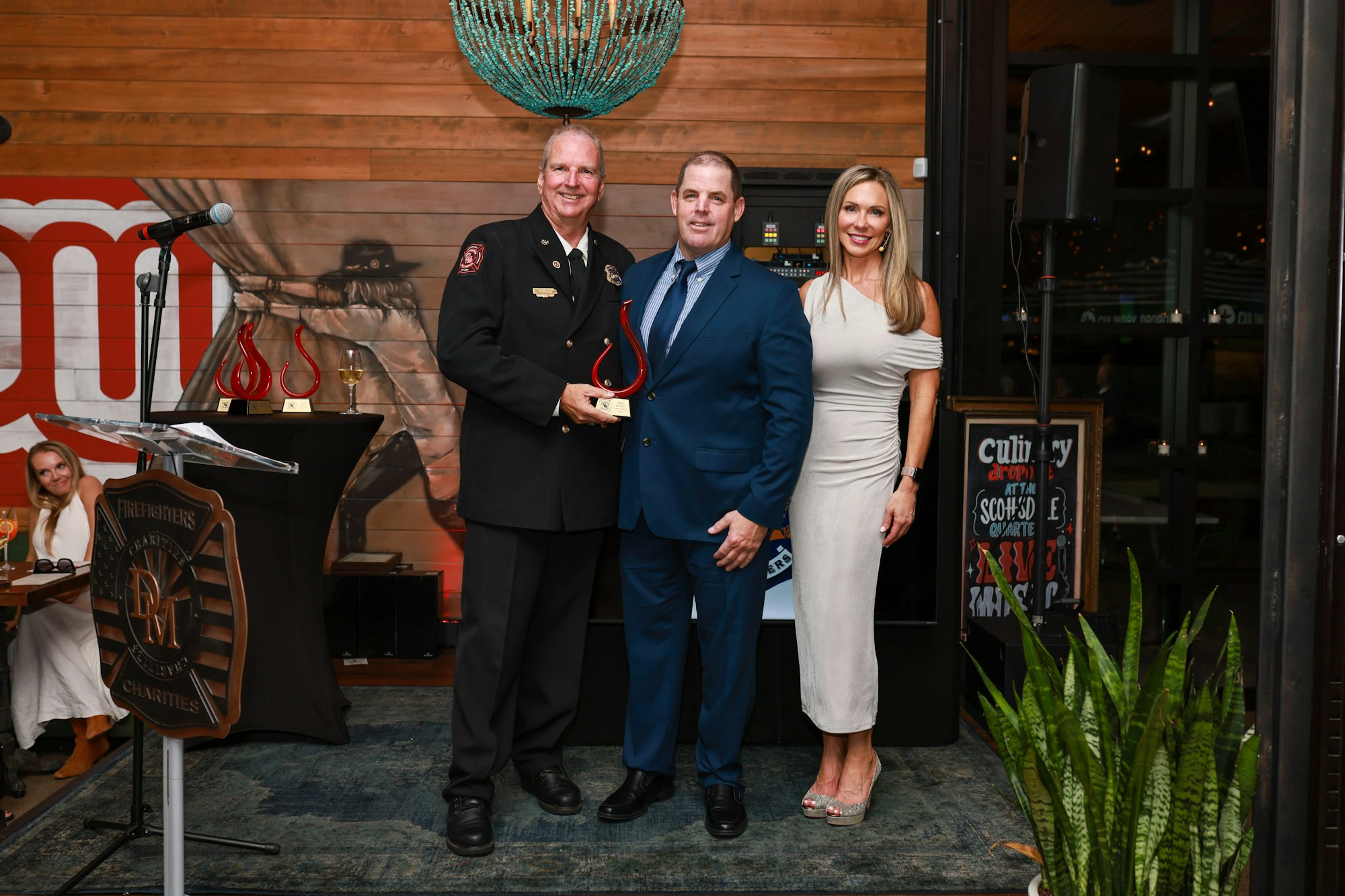 Three people posing with an award, standing in front of a "Firefighters Charities" sign.