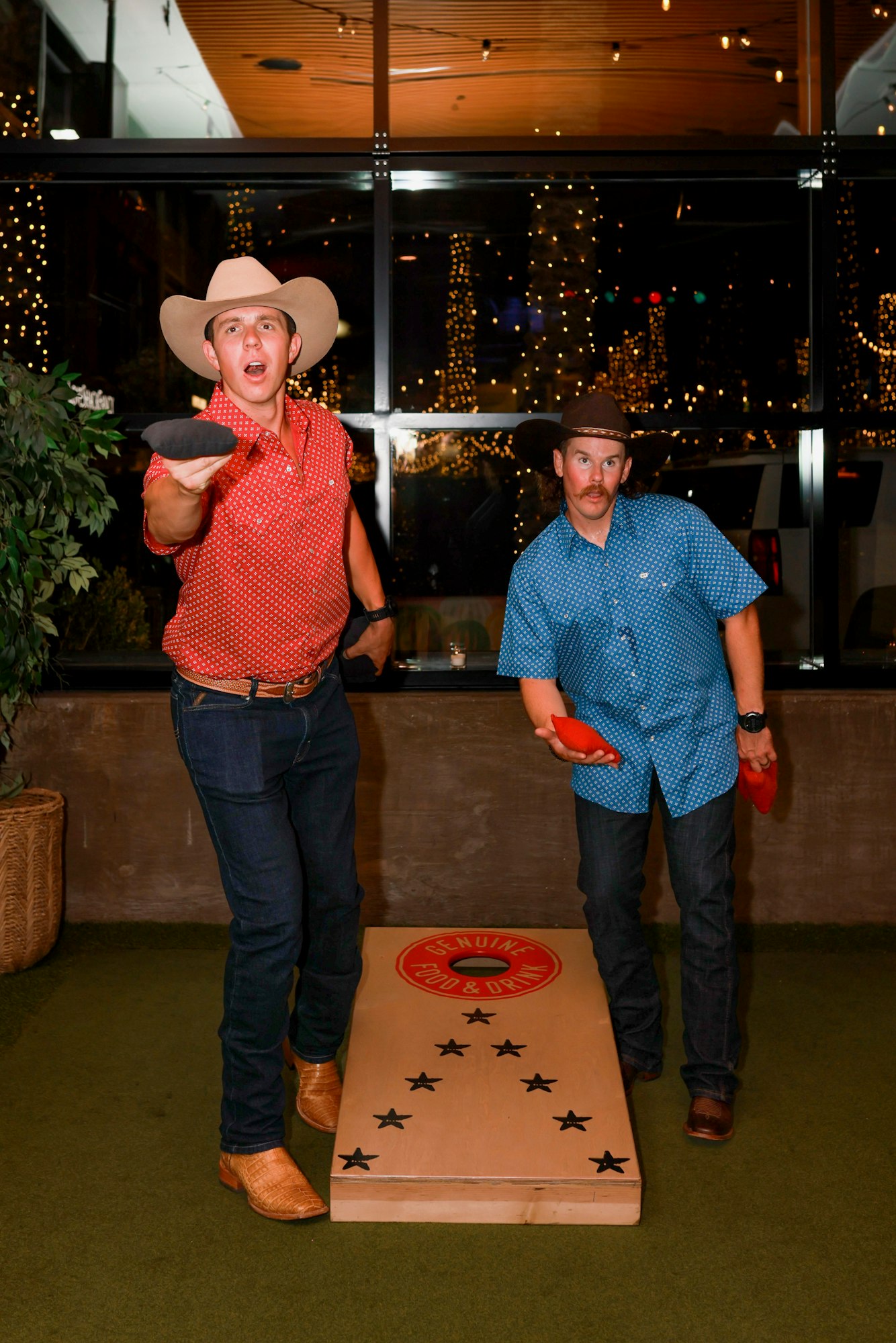 Two men in cowboy hats playing cornhole indoors, one is throwing a beanbag.