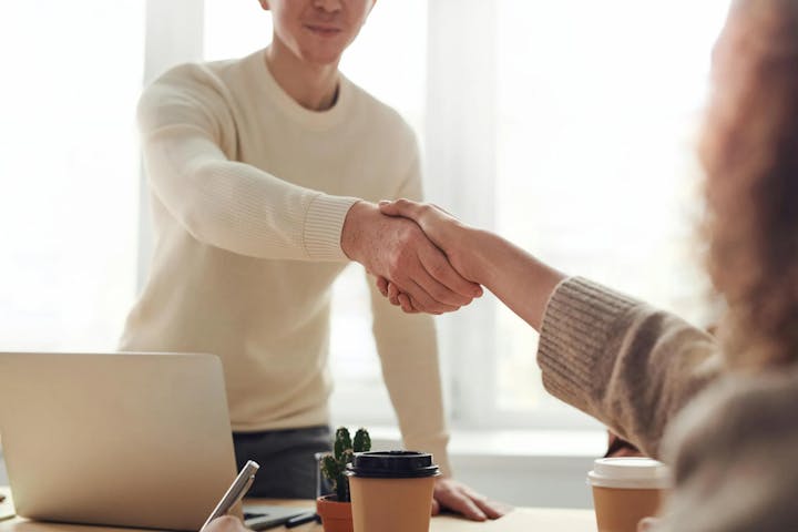 Two people shaking hands over a table with a laptop and coffee cups, suggesting a professional meeting or agreement.