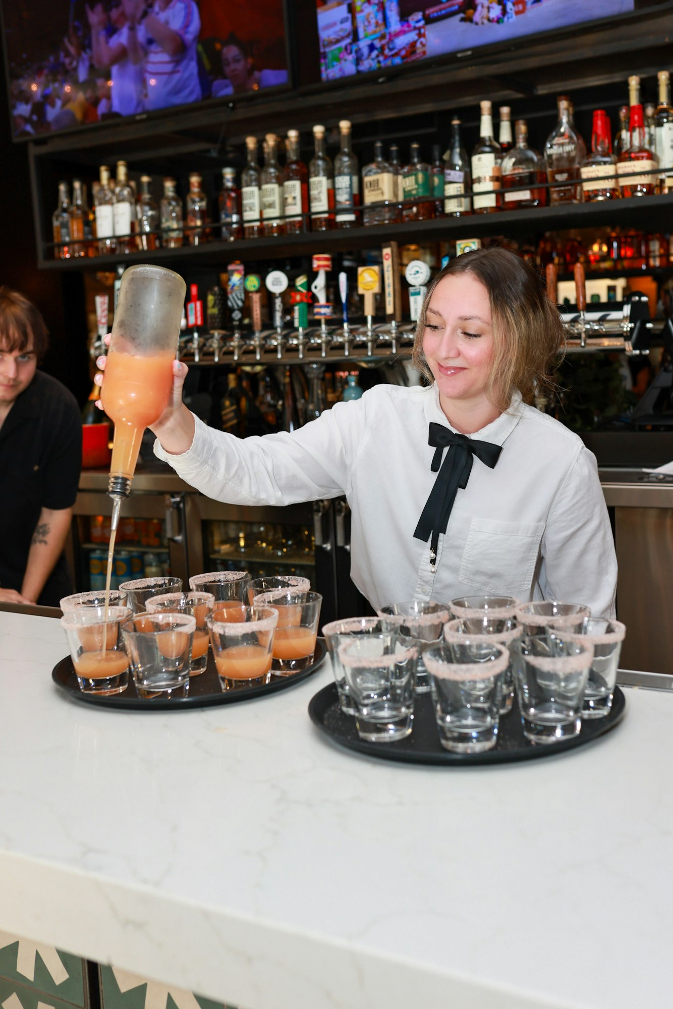 Bartender in white shirt pouring drinks into glasses on trays at a bar.
