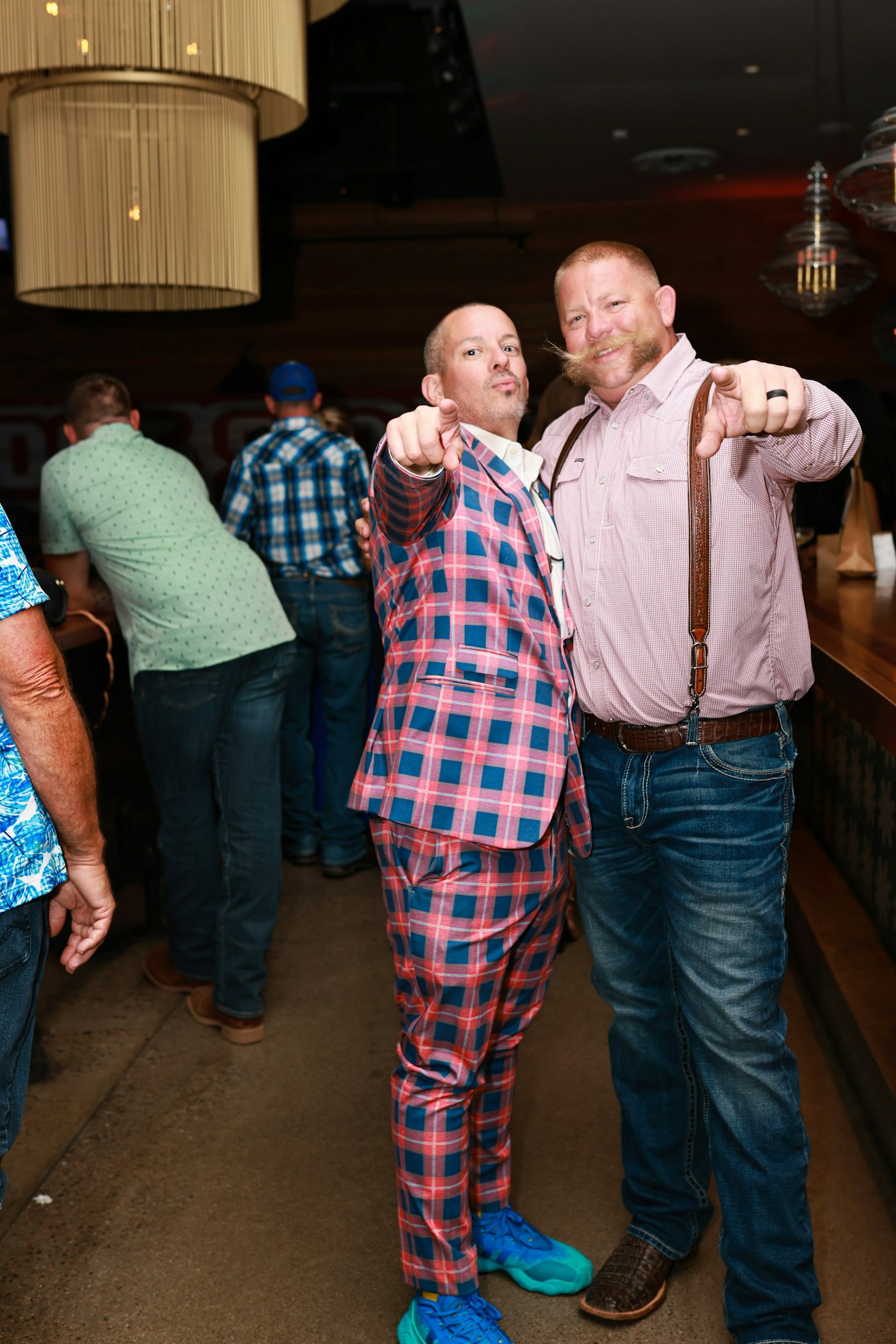 Two men posing and pointing at the camera, one in a plaid suit and blue shoes, the other in jeans and suspenders, in a casual setting.
