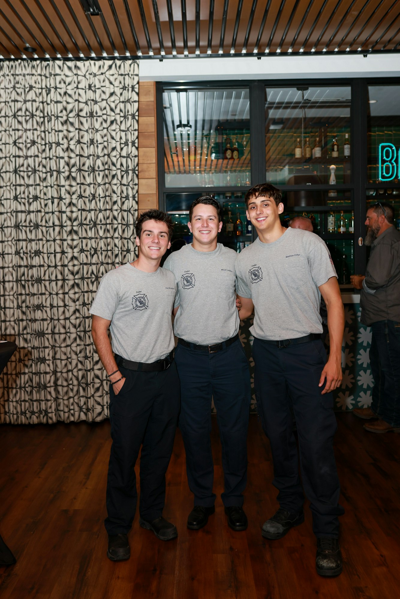 Three men in matching firefighter uniforms smiling at a bar.
