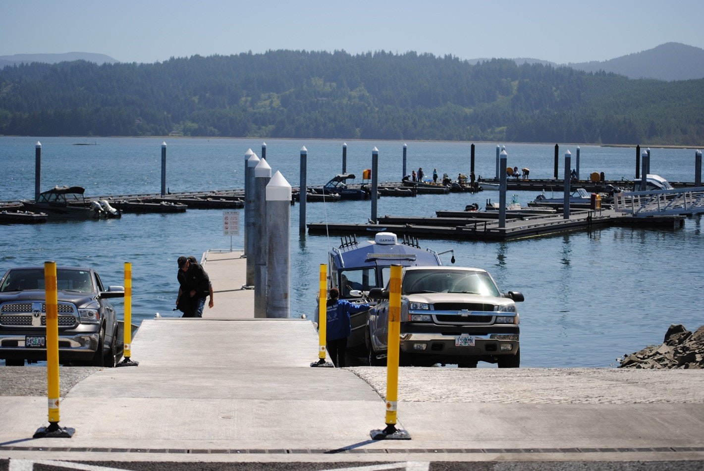 Boat ramp with vehicles, docked boats, and people under a clear sky.