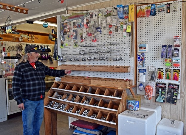 A person in a bait shop pointing at fishing tackle displayed on a pegboard with sorted items underneath.