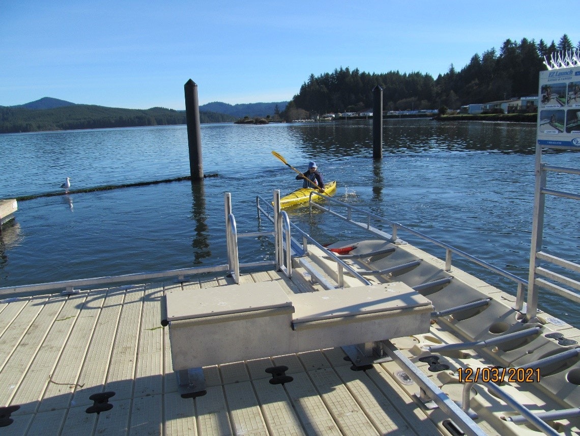 Person in a kayak near a dock with mountain and trees in the background, bright sunny day.