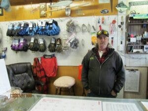 A man standing in a shop with various items displayed on the wall behind him, including footwear and accessories.