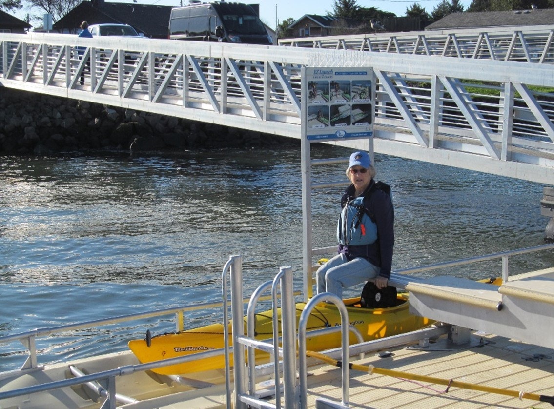 Person on a yellow pedal boat, dock with sign, water, vehicles in the background.