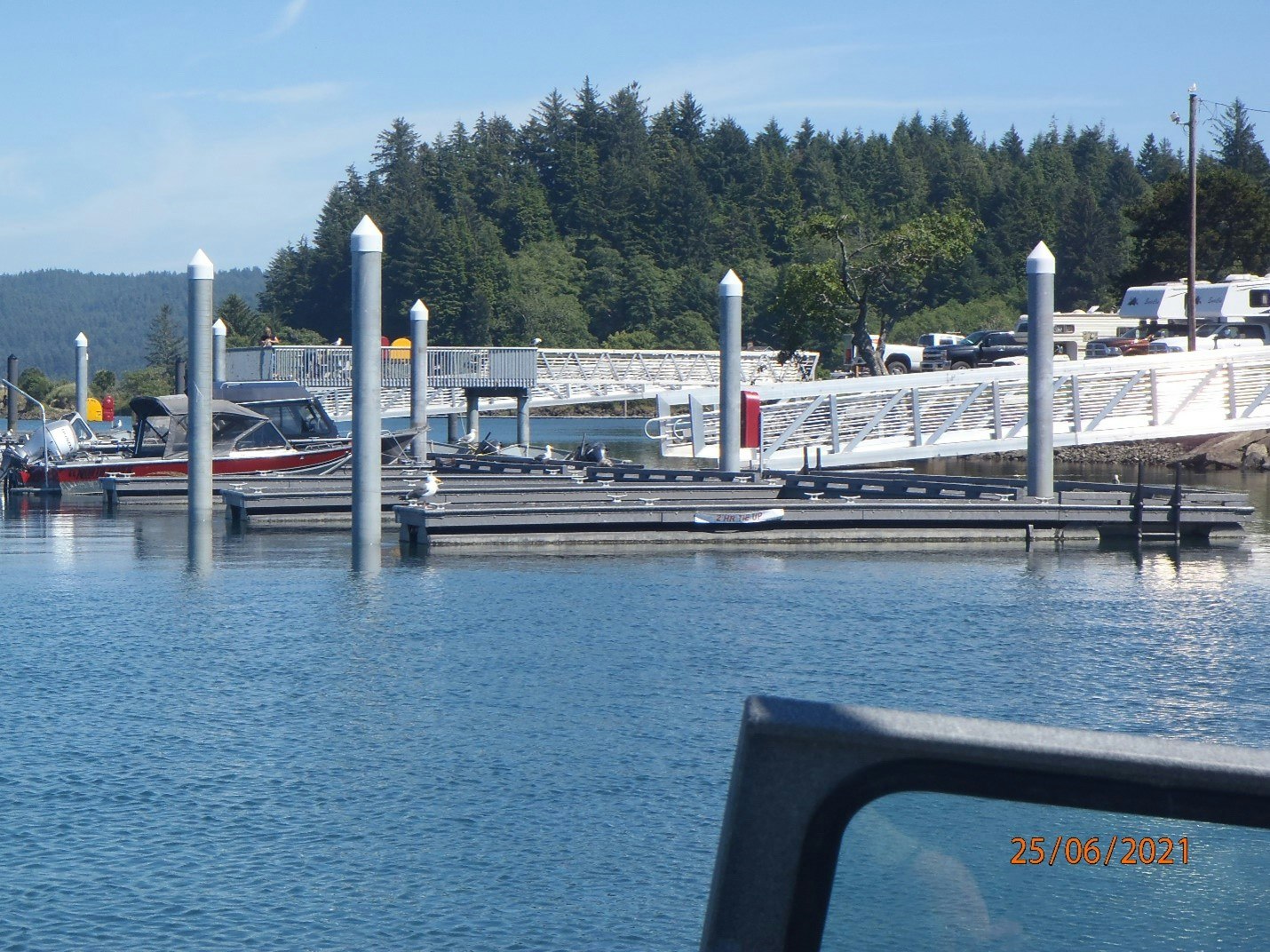 A marina with boats, a forested background, a clear sky, and a partial view of a vehicle window in the foreground.