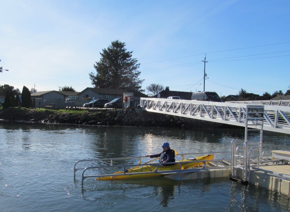 A person kayaking near a dock with a footbridge and houses in the background.