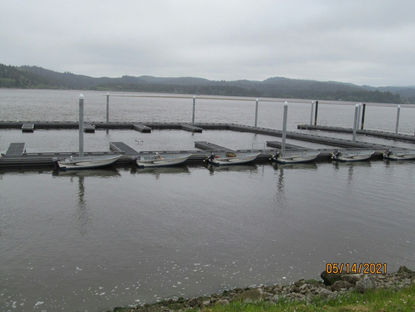 An overcast day at a lake with empty docks and rowboats tied up, with calm water and a hilly backdrop.