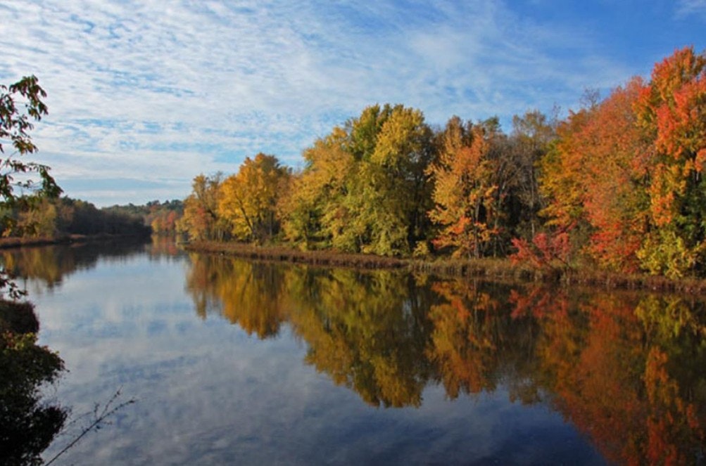A serene river scene adorned with vibrant autumn foliage reflecting on the water under a blue sky with scattered clouds.