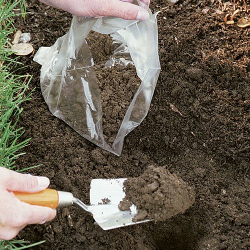 A person is using a hand trowel to add soil from a plastic bag into a dug hole in the ground.
