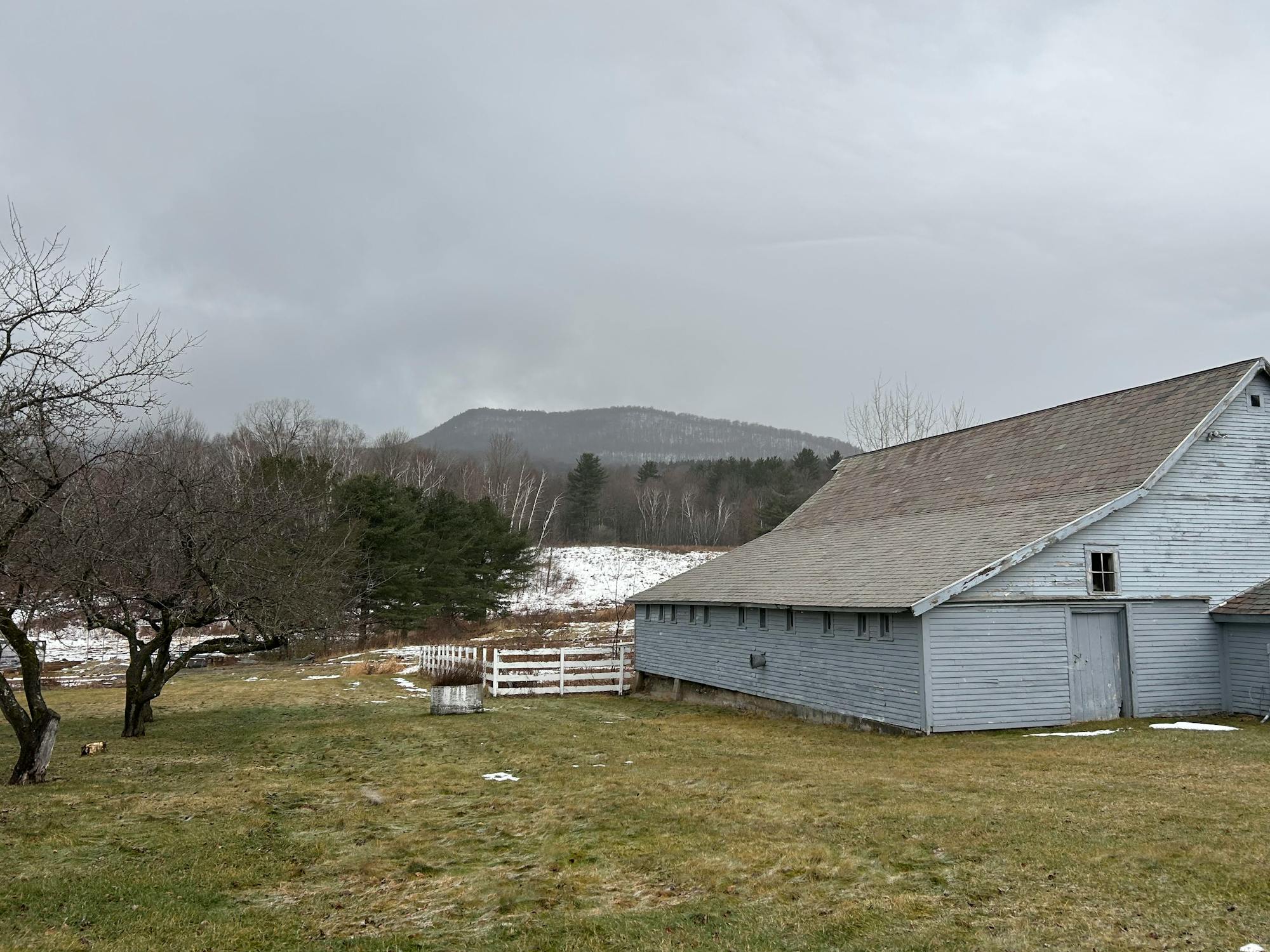 A gray barn is set against a cloudy sky and distant mountains, surrounded by grassy fields and sparse trees.