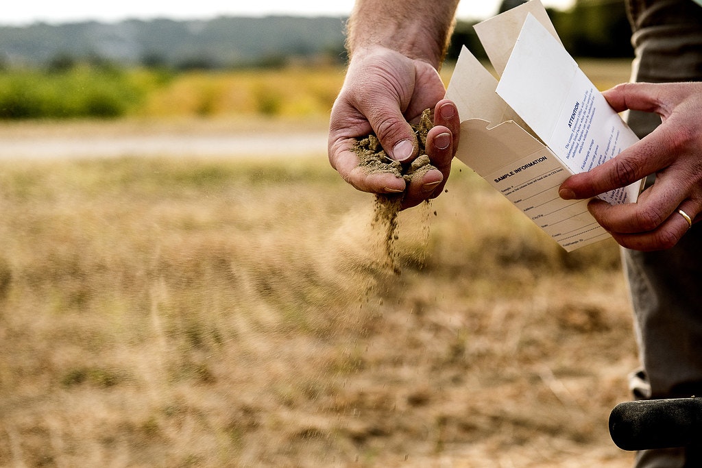 A person is holding a packet and sprinkling soil or sand from their hand, possibly for sampling purposes in a field.