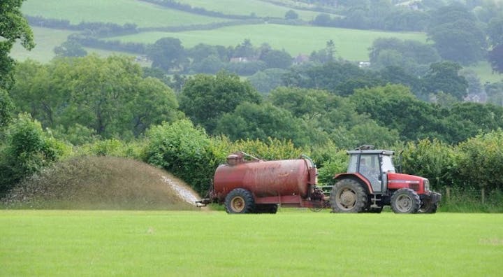 A red tractor is spreading liquid fertilizer on a green field, surrounded by trees and hills.