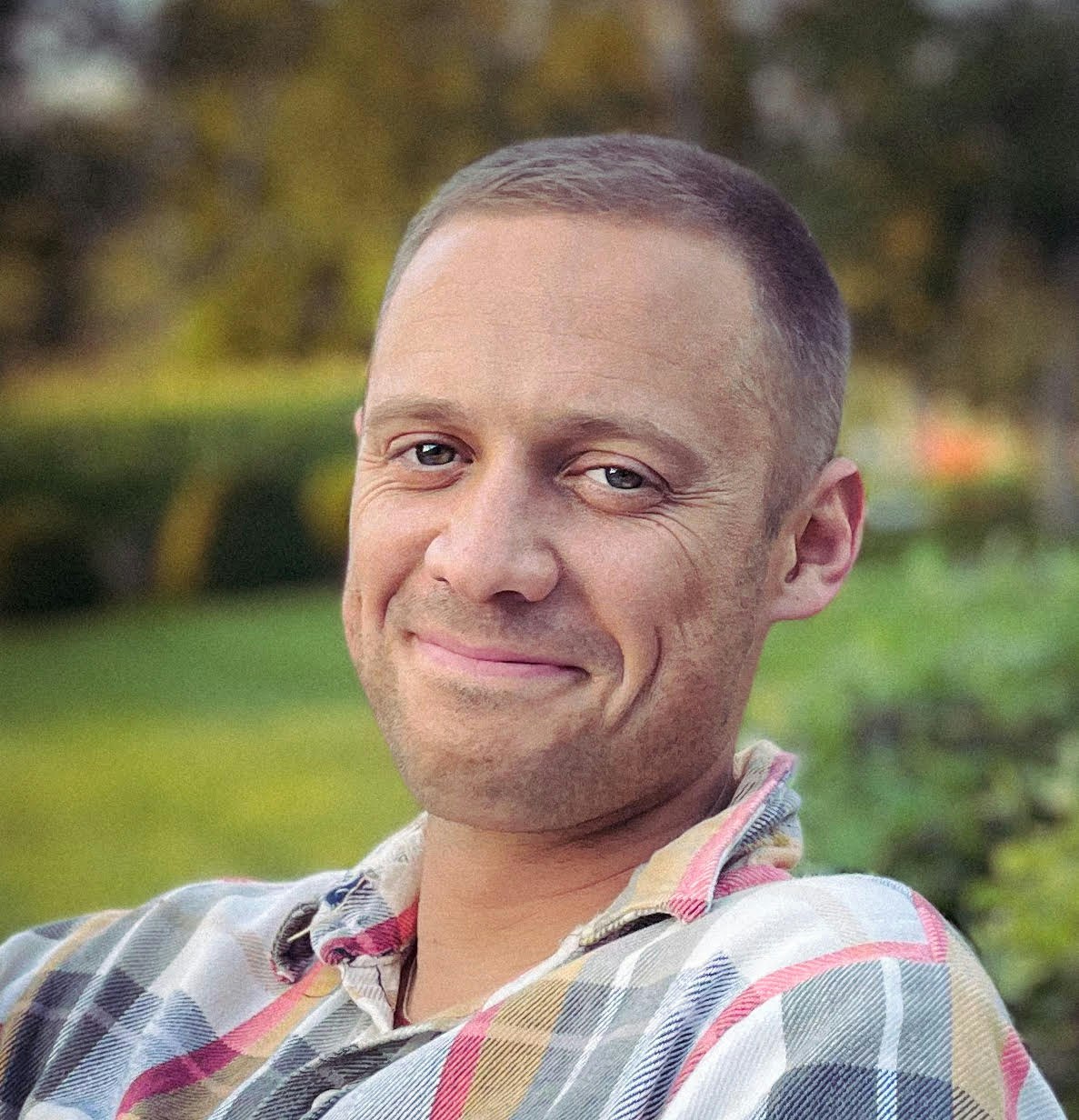 A smiling person sits outdoors, wearing a plaid shirt, with greenery in the background. The atmosphere is calm and pleasant.