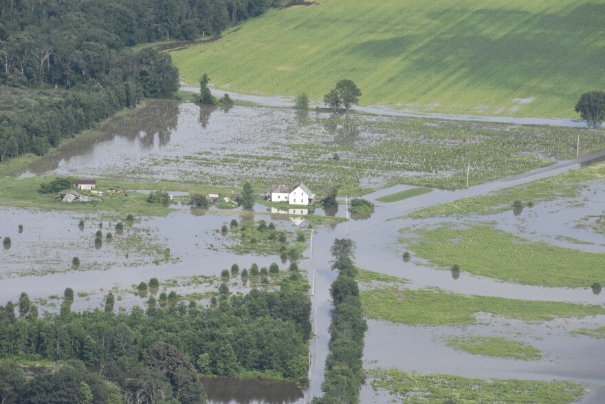 Aerial view of a flooded area with a house, surrounded by water and green fields.