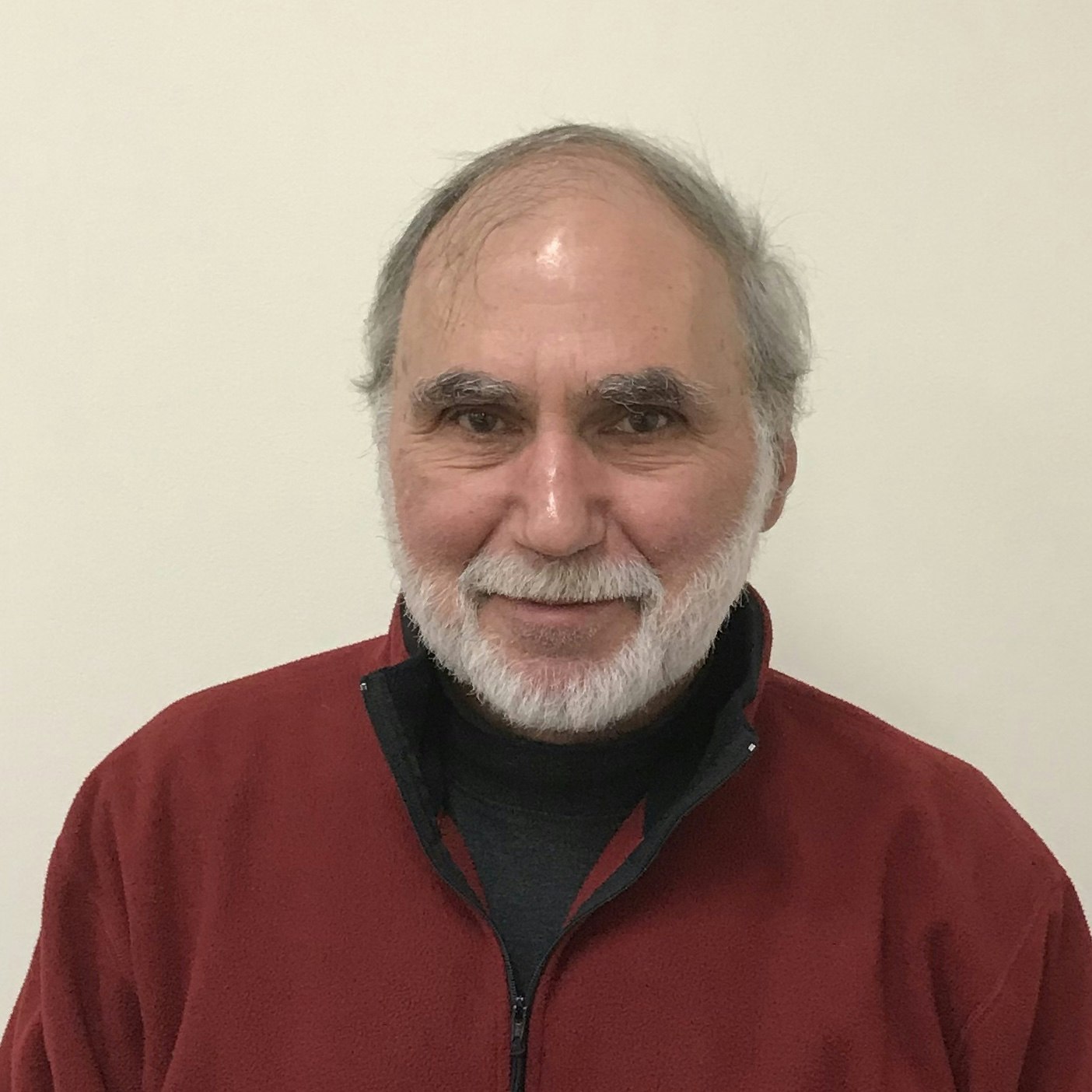 A smiling older man with a beard is wearing a red jacket against a plain background.