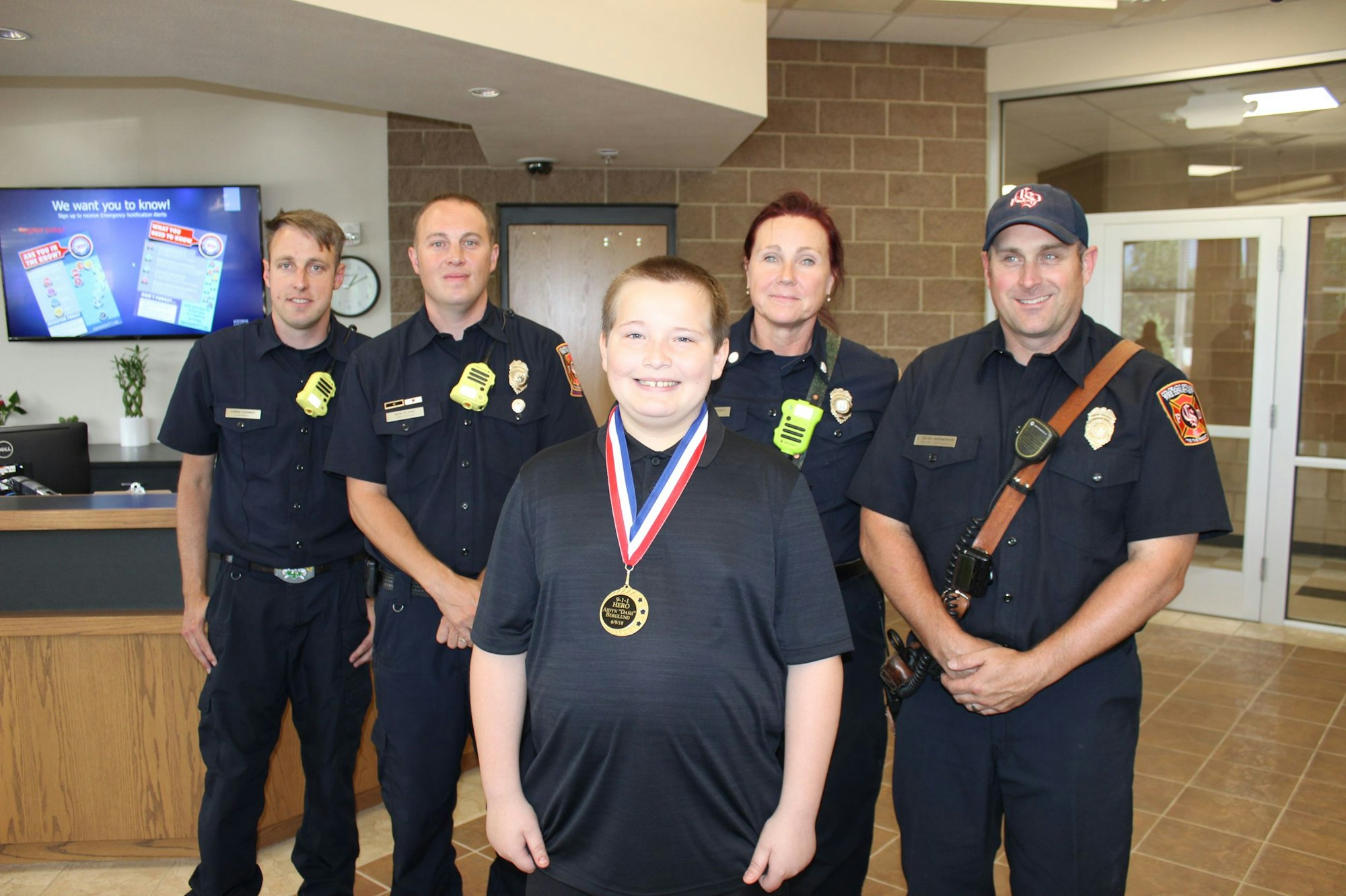 A smiling child with a medal stands in front of four uniformed firefighters indoors.