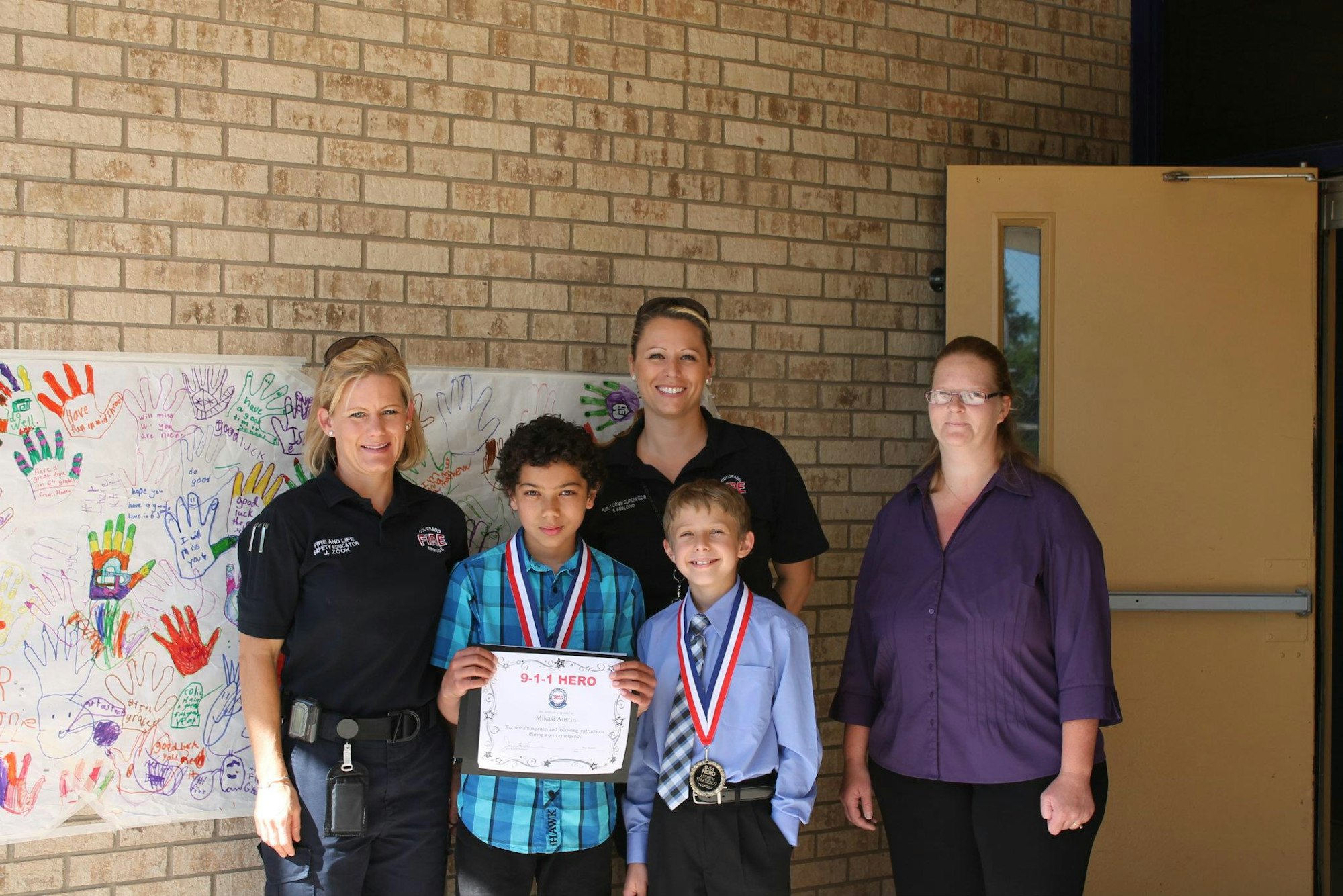 Two adults, two children with medals, and a woman stand by a wall with colorful hand drawings.