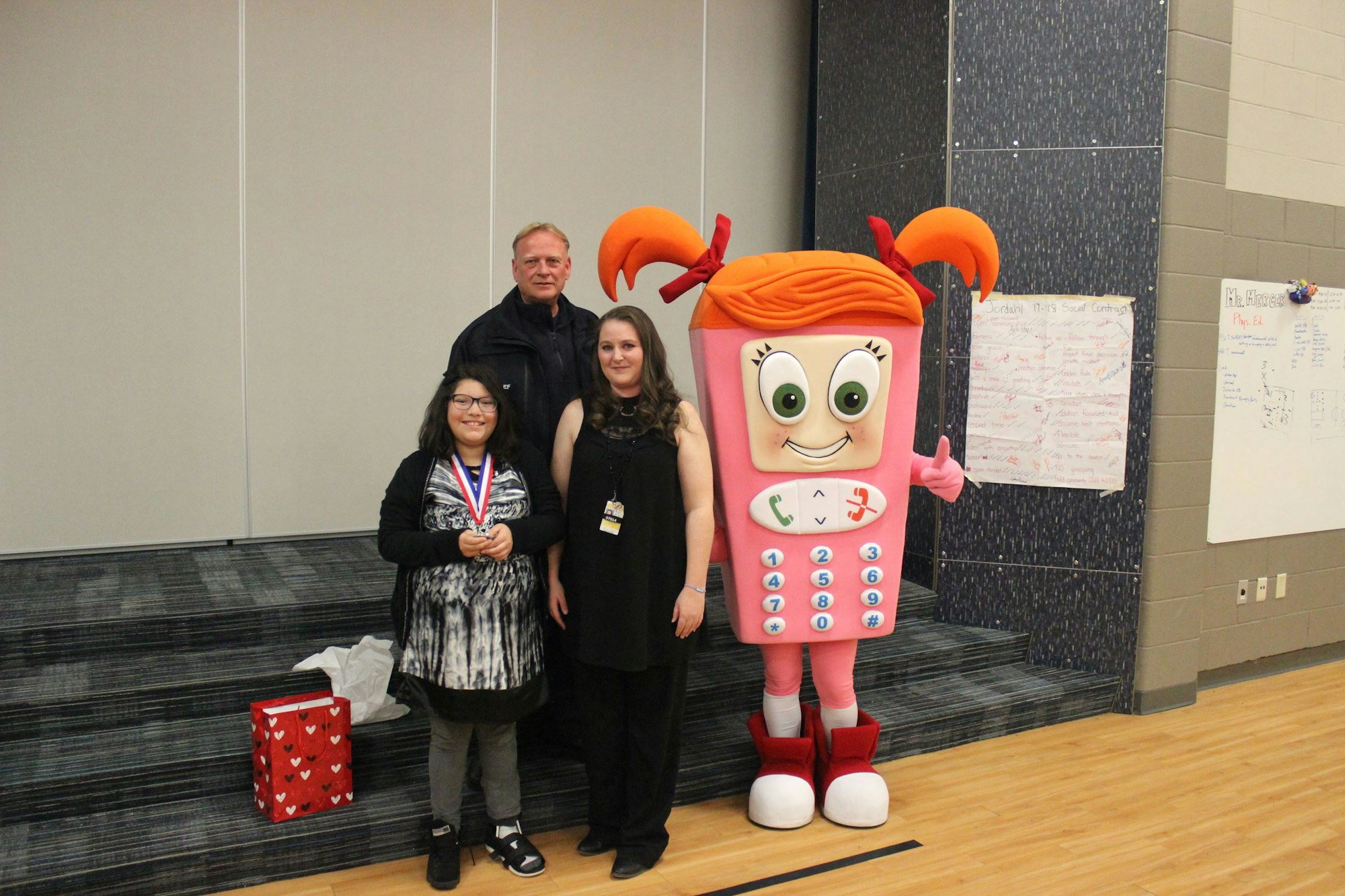 Four people, including one in a phone costume, stand together; one holds a medal.