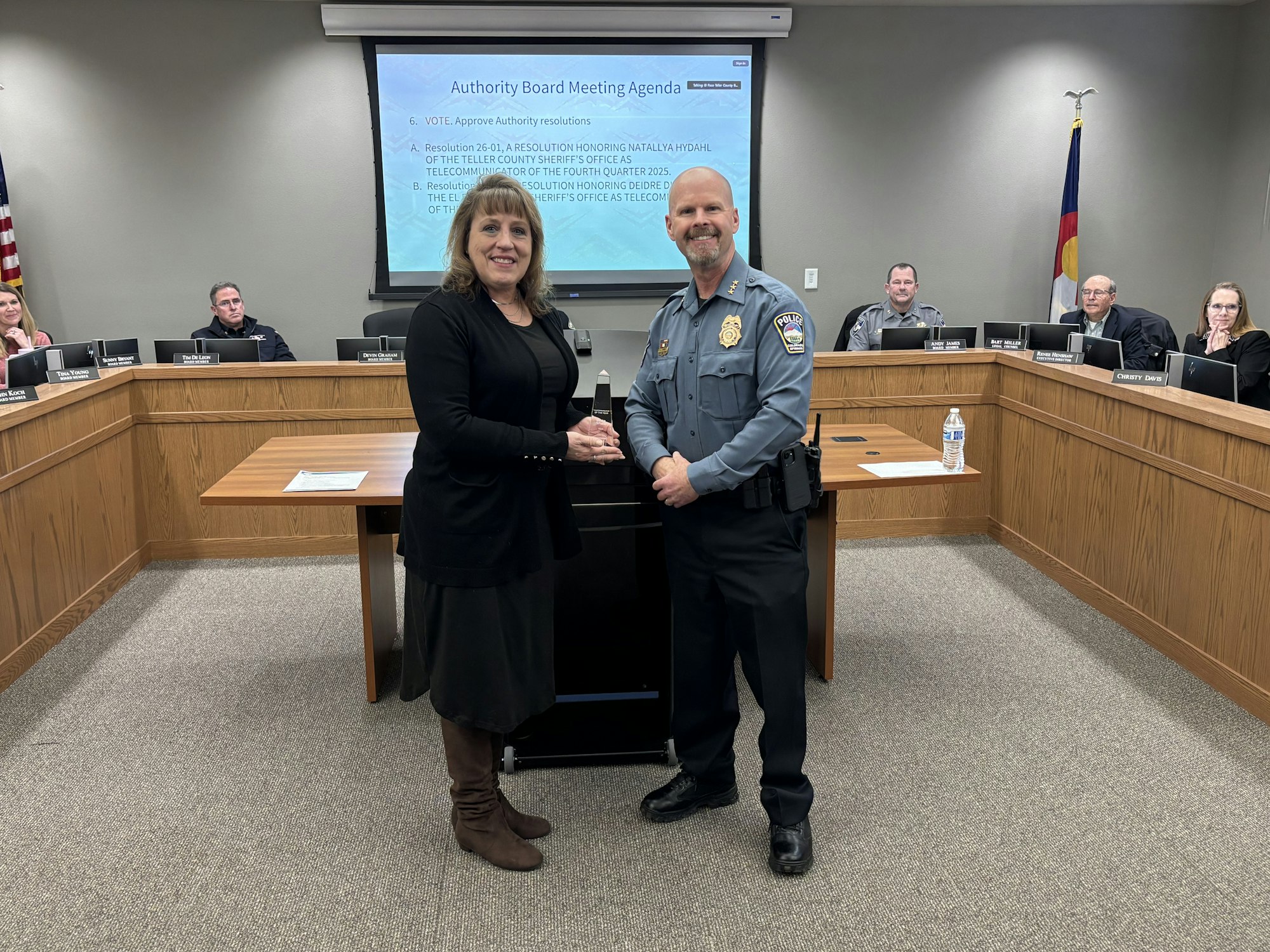 A smiling woman holds an award with a police officer in a board meeting room, while others watch from the background.