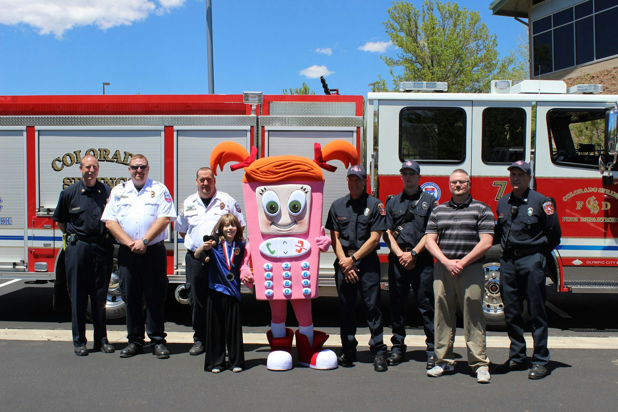 A group of firefighters, a child, and a mascot are posing in front of a fire truck.