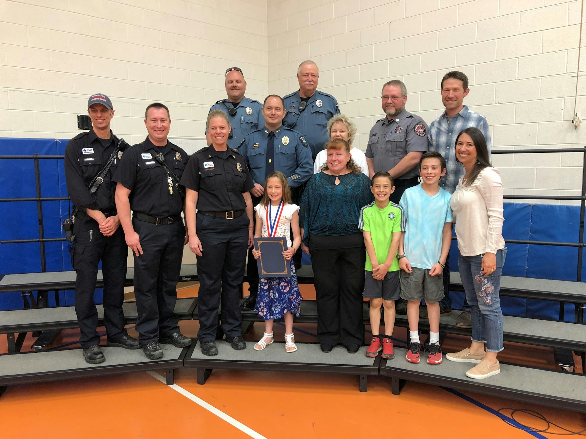 A group of people, including police officers and civilians, pose for a photo. A young girl in front holds a plaque and wears a medal.