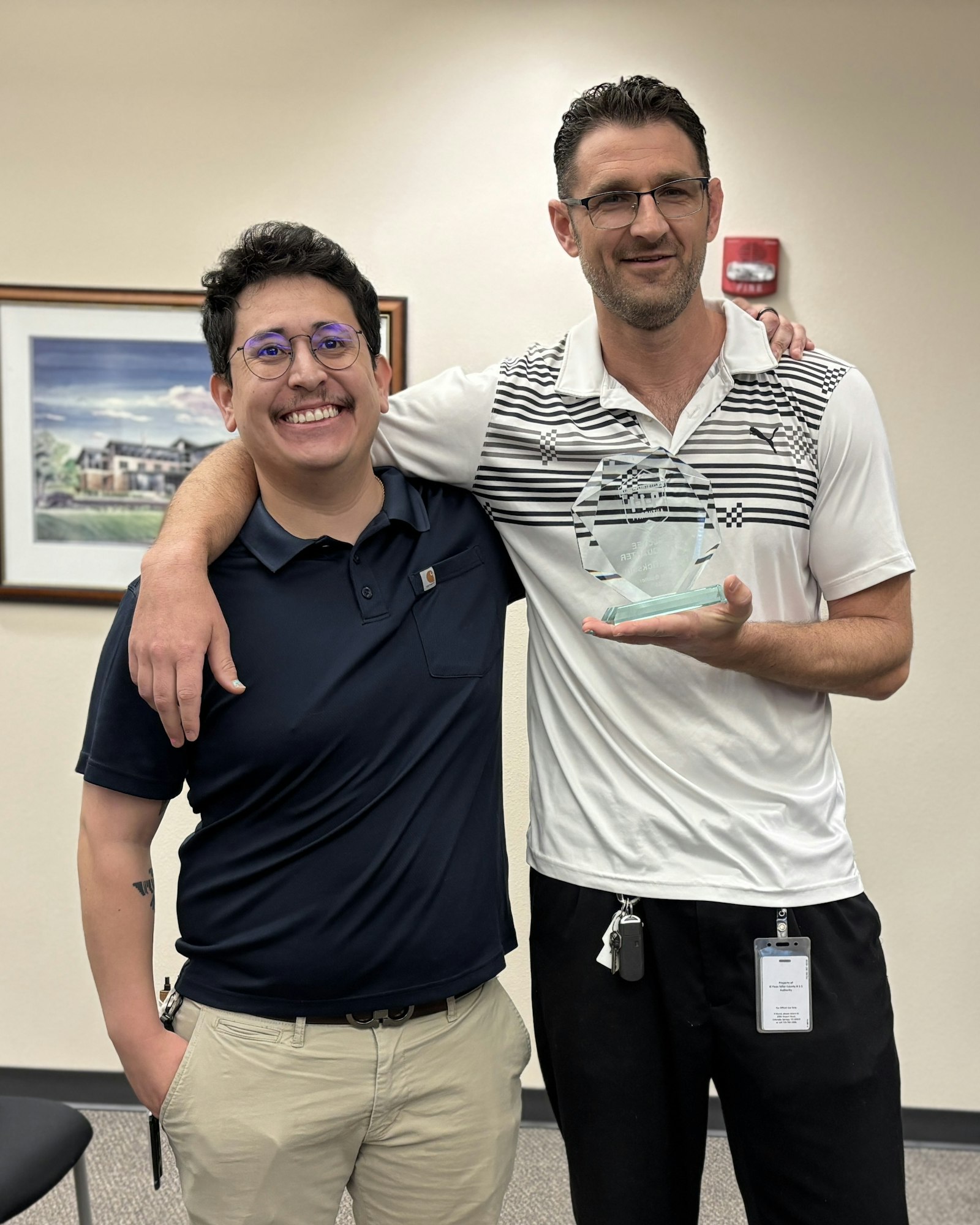 Two people smiling, one holding a glass award, standing in an office setting.