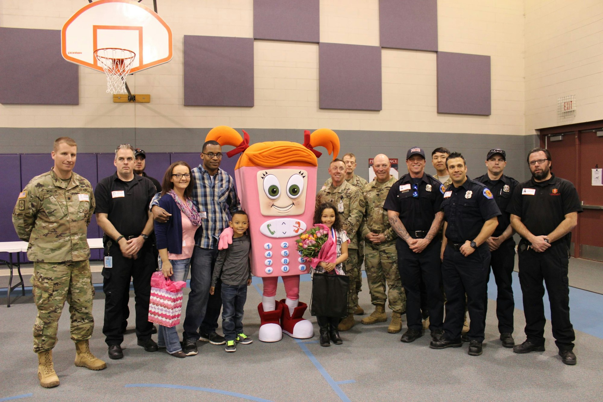A group poses with a phone mascot in a gym. Adults and children stand with military and emergency service personnel.