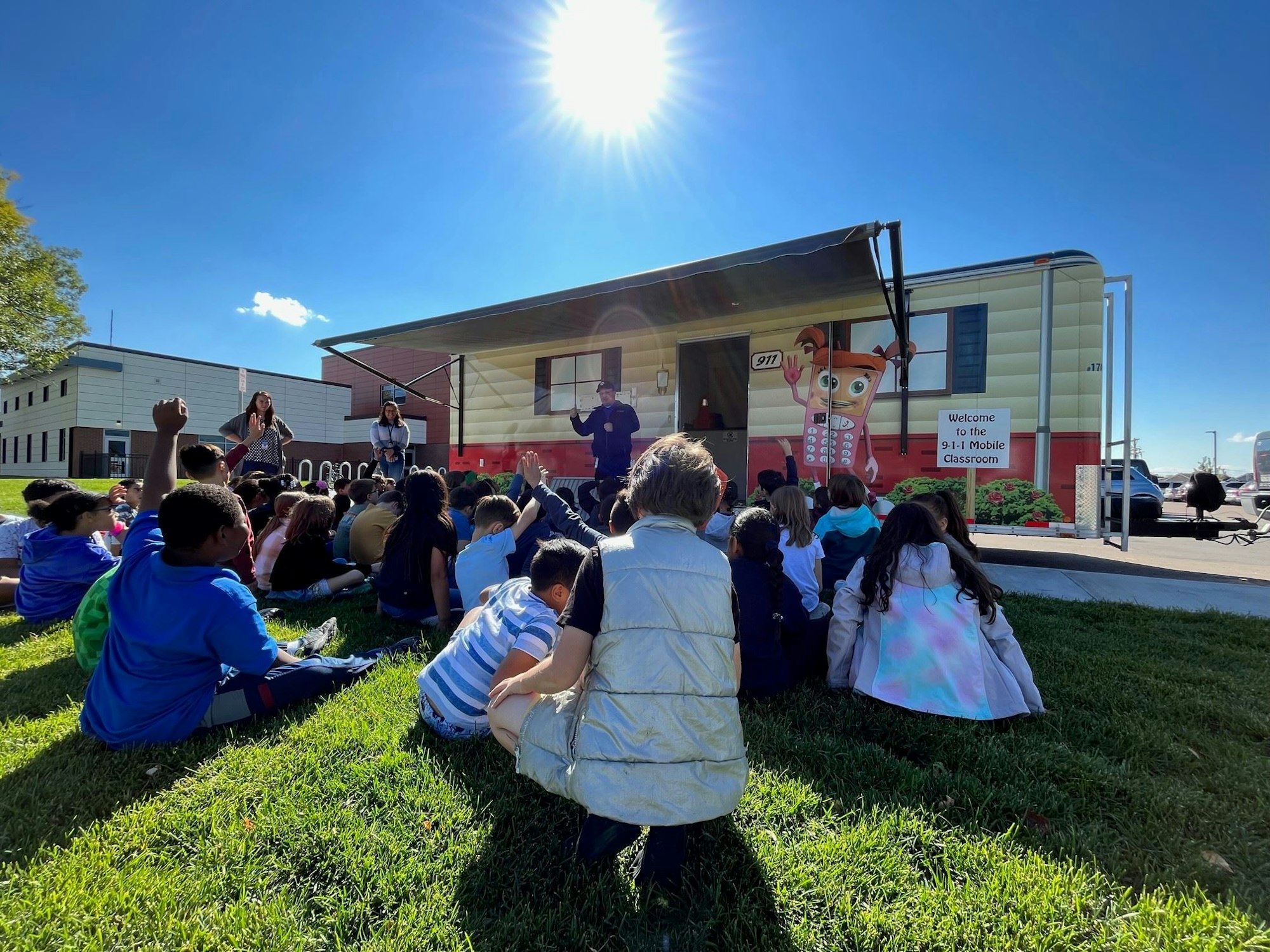 A group of children sits on grass near a "911 Mobile Classroom," listening to instructors under a sunny sky.