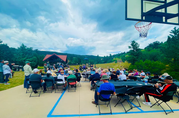 Outdoor gathering with people sitting on chairs around tables. A basketball hoop is visible, set against a backdrop of trees and a cloudy sky.