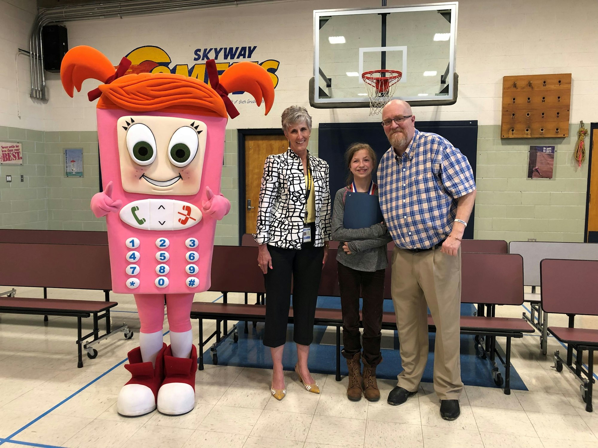 Three people smiling with a large phone mascot in a gymnasium.