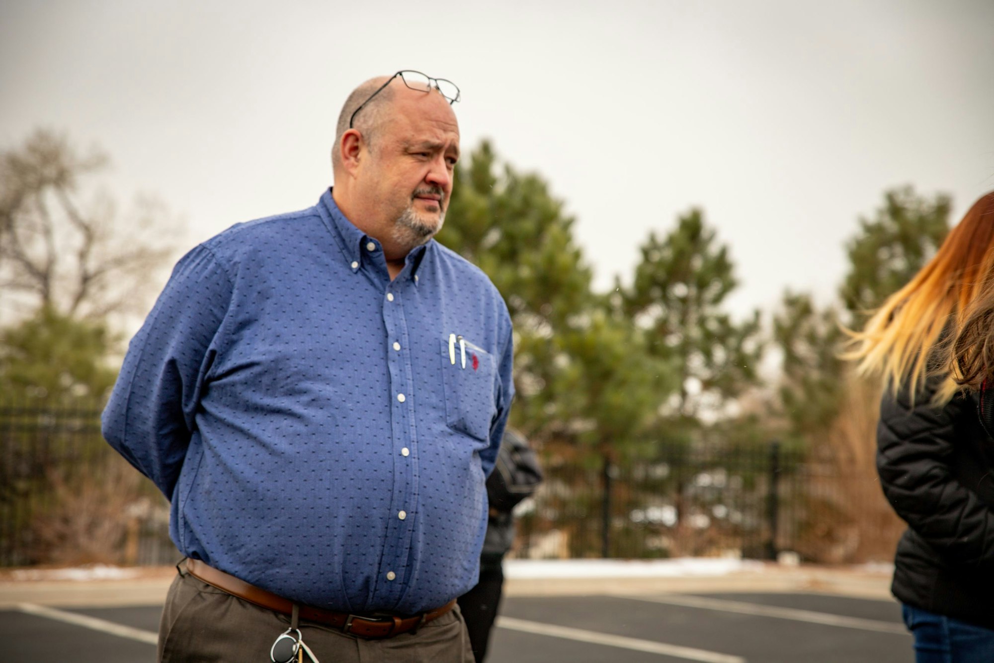 A person in a blue shirt stands outdoors, looking to the side with trees in the background.