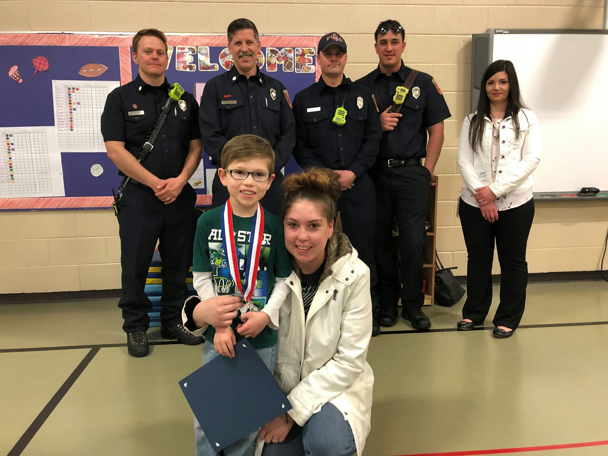 A child with a medal and certificate poses with firefighters and others in a gymnasium-like setting.