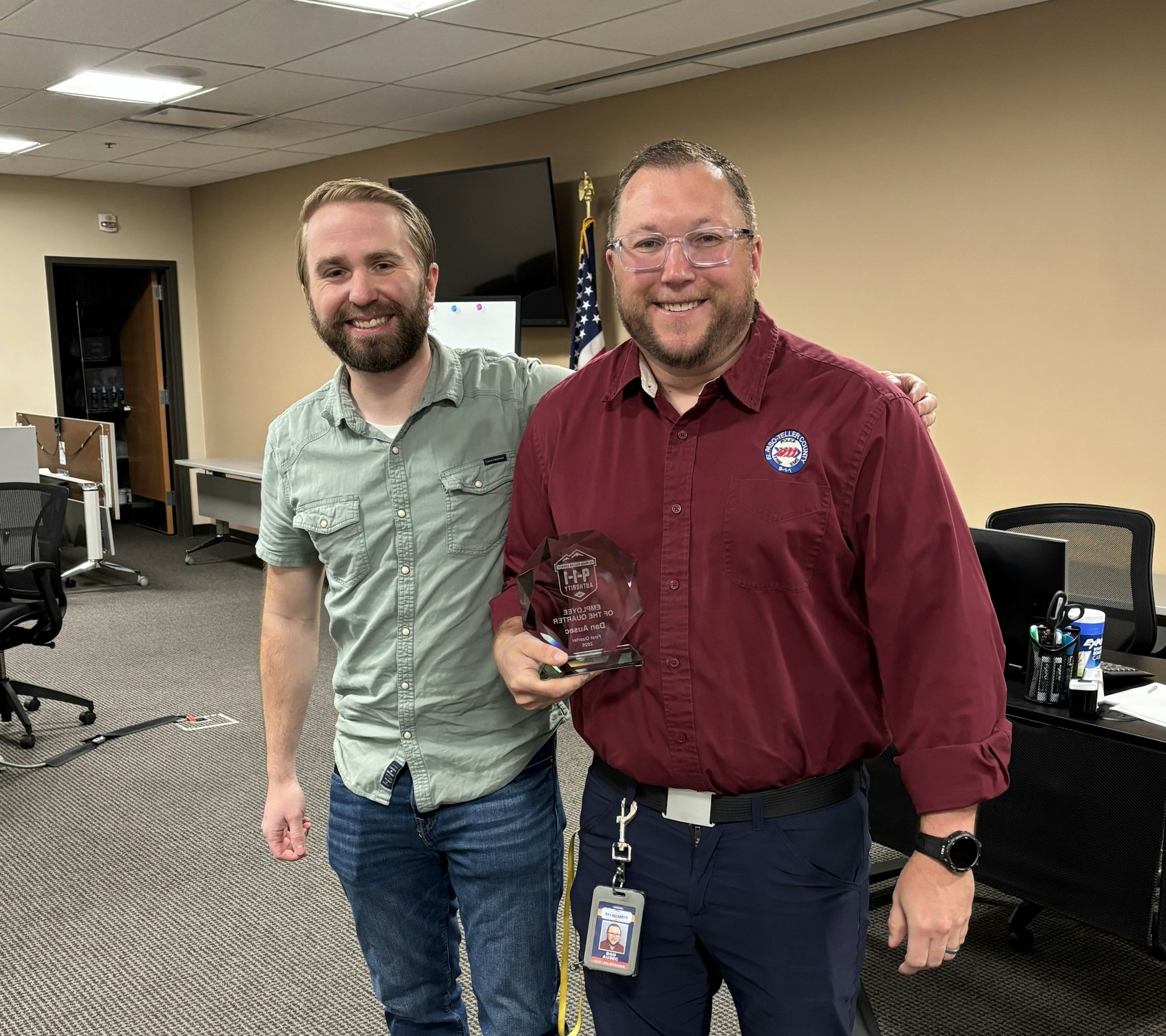 Two men stand together, one holding an award while smiling. The setting appears to be an office or conference room.