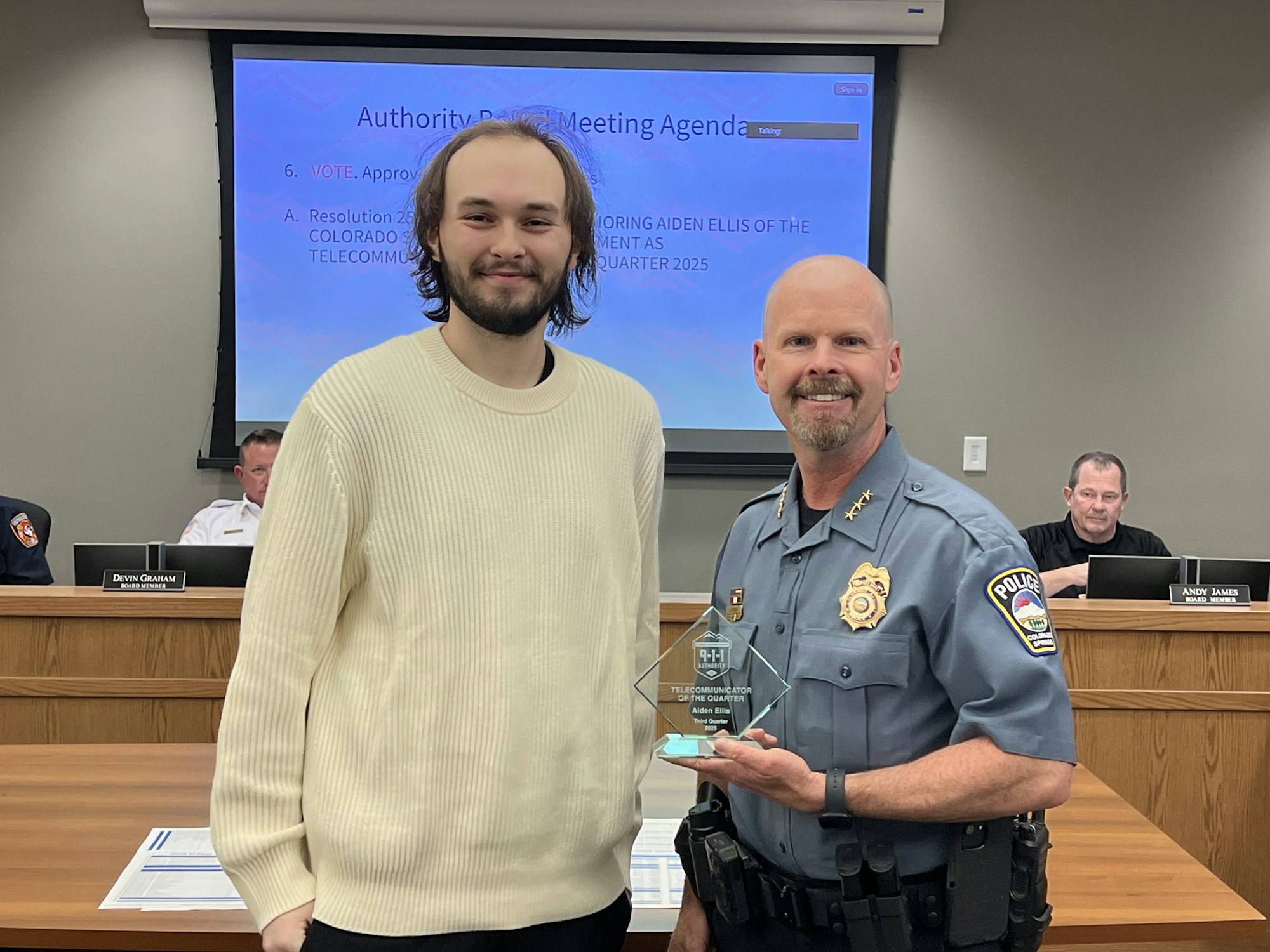Two men are standing; one holds an award. A presentation is displayed in the background.