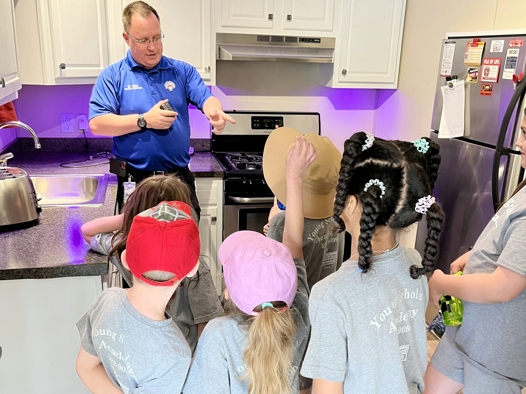 A man in a blue shirt talks to a group of children in gray shirts inside a kitchen.