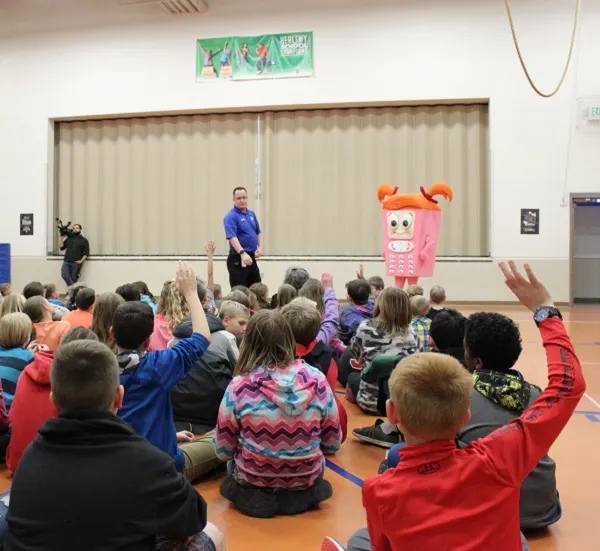 A group of children seated in a gymnasium, with a person in a pink cartoon phone costume and an adult presenter at the front.