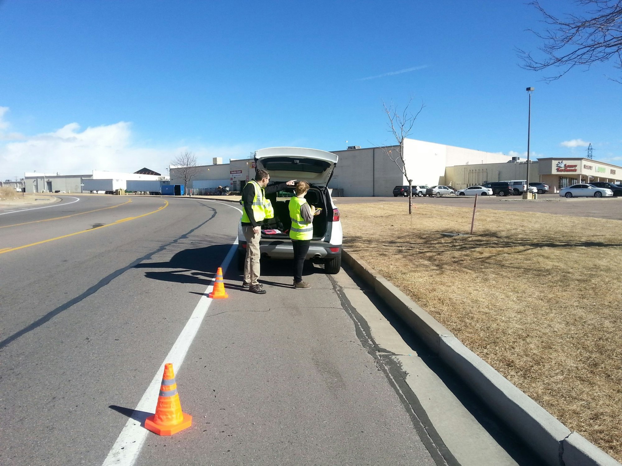 Two people wearing safety vests stand by an open vehicle trunk on the roadside, near traffic cones.