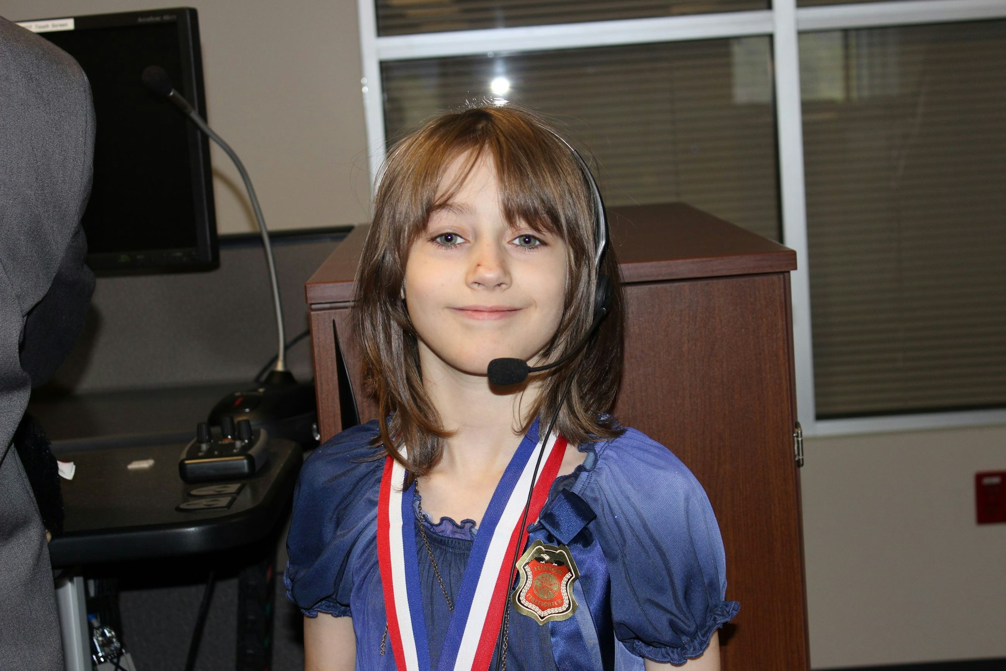 A child wearing a headset and medals, standing indoors with a desk and computer in the background.